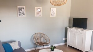 A living room with a grey sofa, a wicker chair, a white cabinet with a TV, and three framed pictures on the wall.
