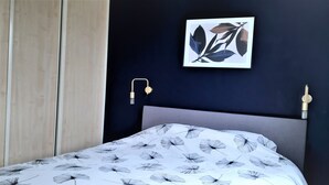 A bedroom with a bed featuring a floral-patterned bedspread, a dark wall, a framed artwork, and a bedside lamp.