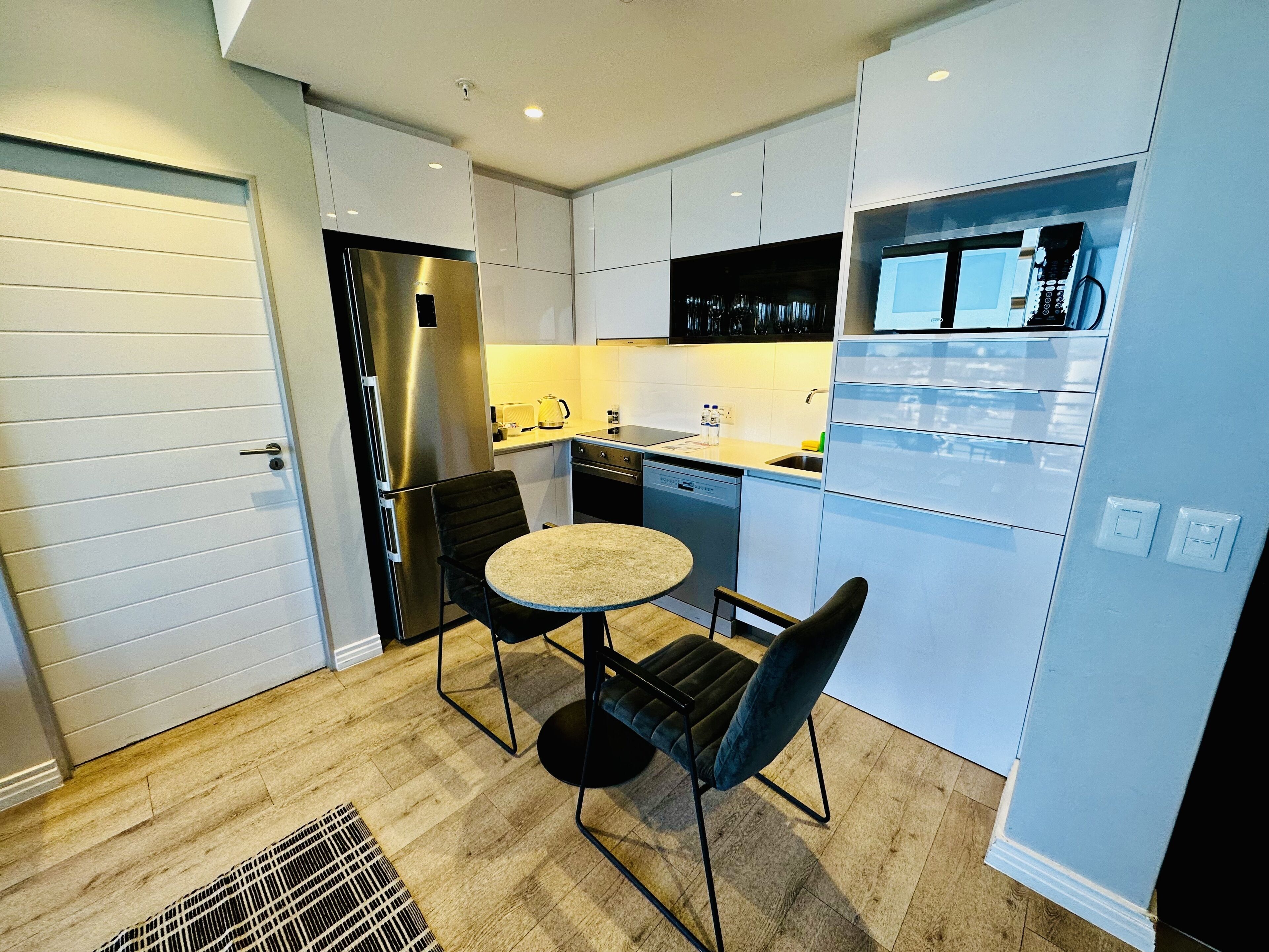 Una cocina compacta con muebles blancos, un refrigerador de acero inoxidable, un lavavajillas y una pequeña mesa de comedor con dos sillas.