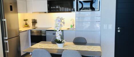 A modern kitchen with white cabinetry, a built-in microwave, and a stainless steel refrigerator. There is a dining table with two chairs and a potted orchid.