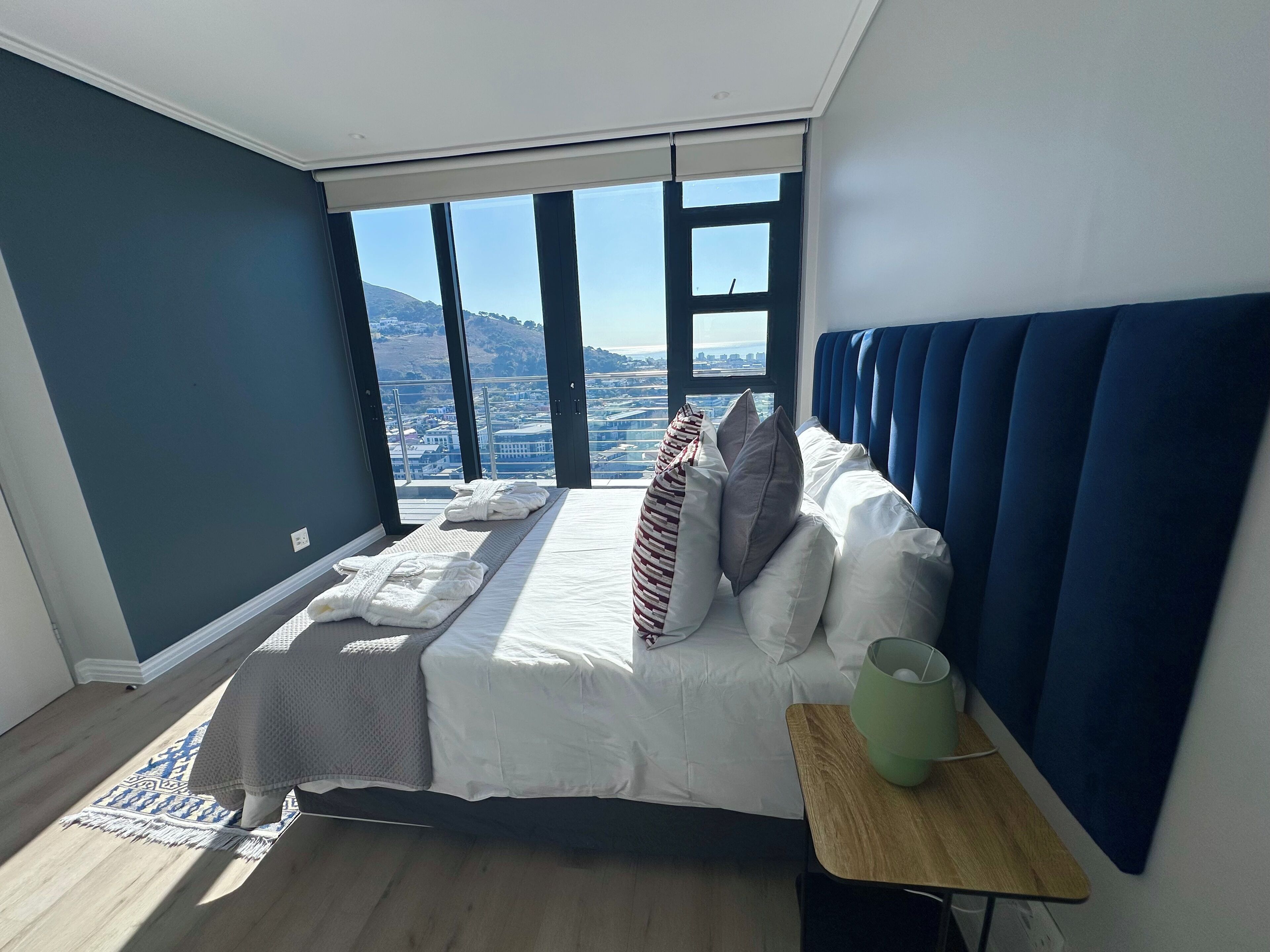 Un dormitorio con un ventanal grande, una cama con ropa de cama blanca, cabecera azul y una mesita de noche pequeña con una lámpara.