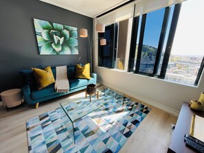 A modern living room with a teal sofa, patterned rug, and a large window offering a city view. - Urban Elephant 16 on Bree (Cape Town)