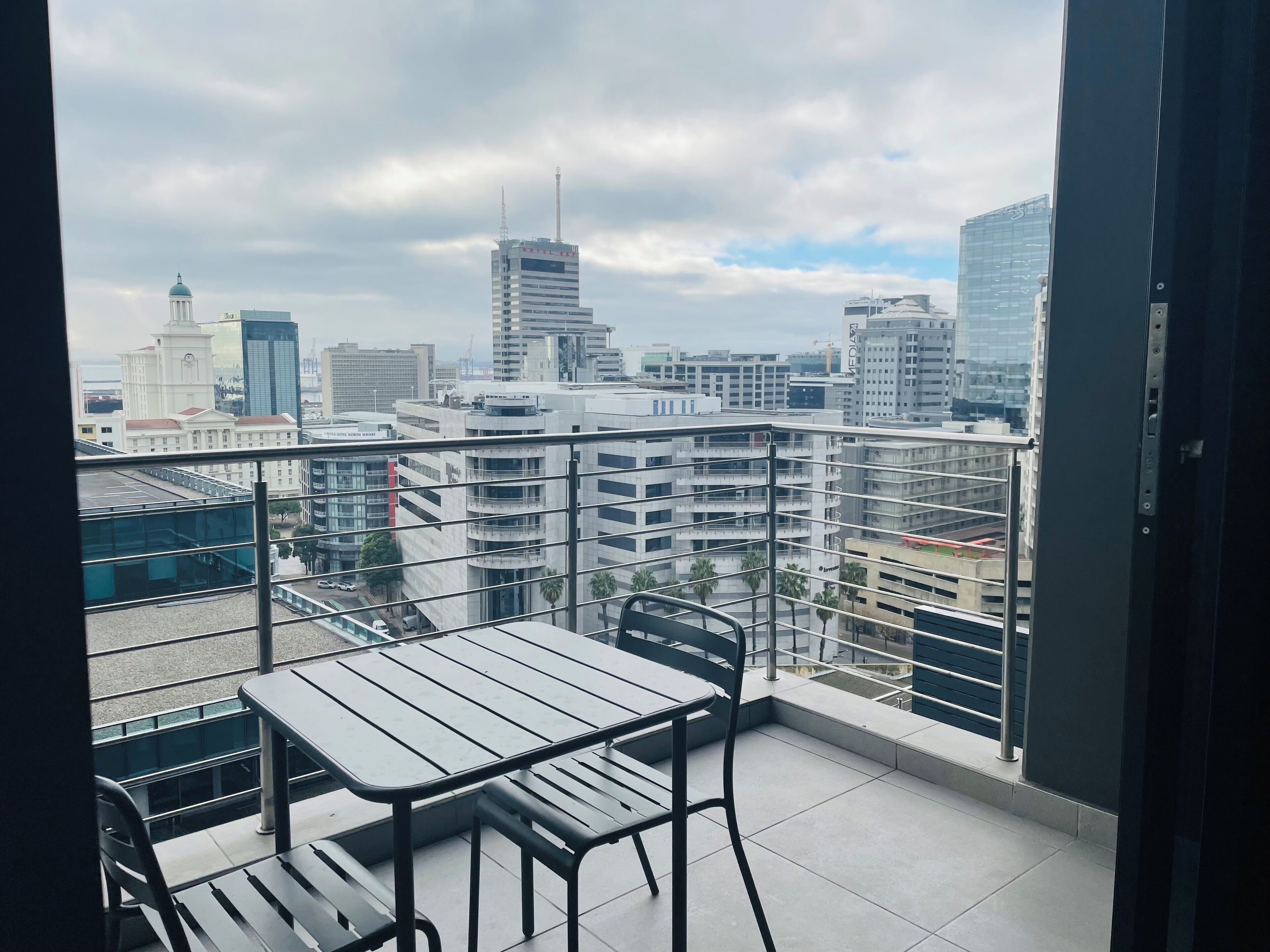 A balcony with a table and chairs, overlooking a cityscape with modern buildings and a clock tower.