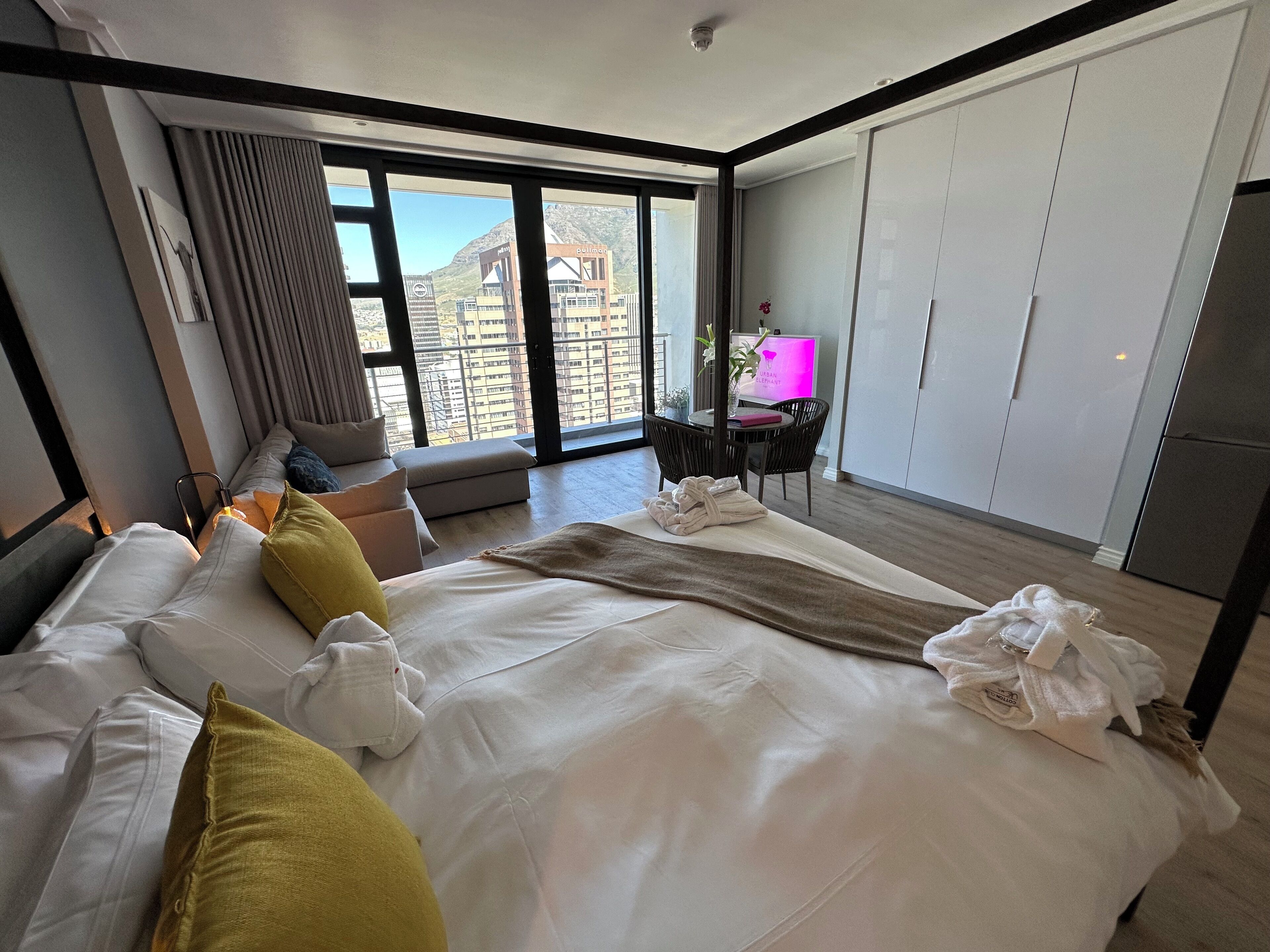 Una habitación de hotel moderna con una cama grande, una zona de estar con cojines, un escritorio con una computadora y vistas a un paisaje urbano a través de amplias ventanas.