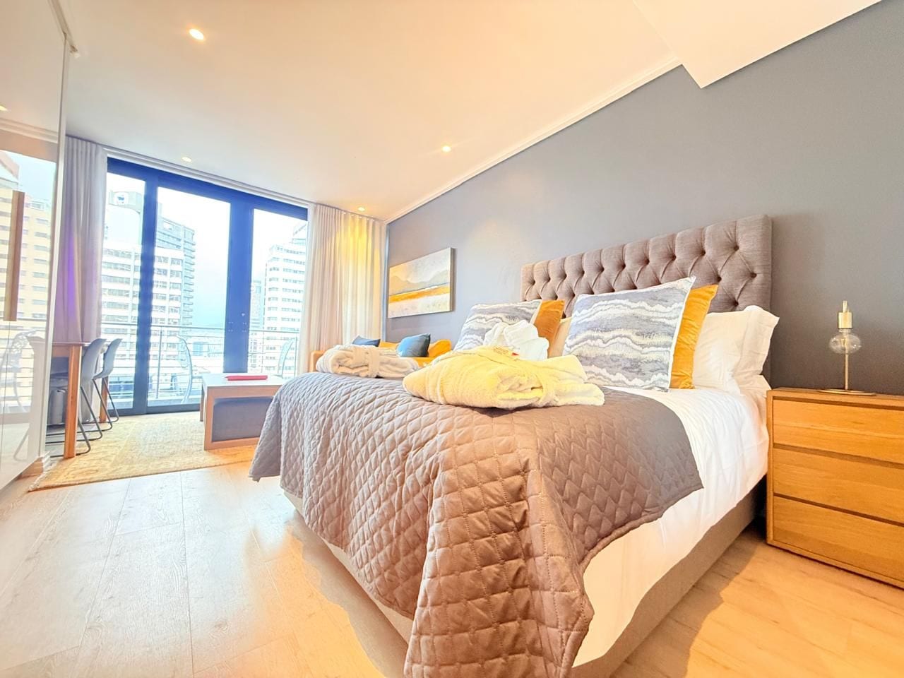 A modern bedroom with a large bed, a bedside table, and a view of the city through large windows.