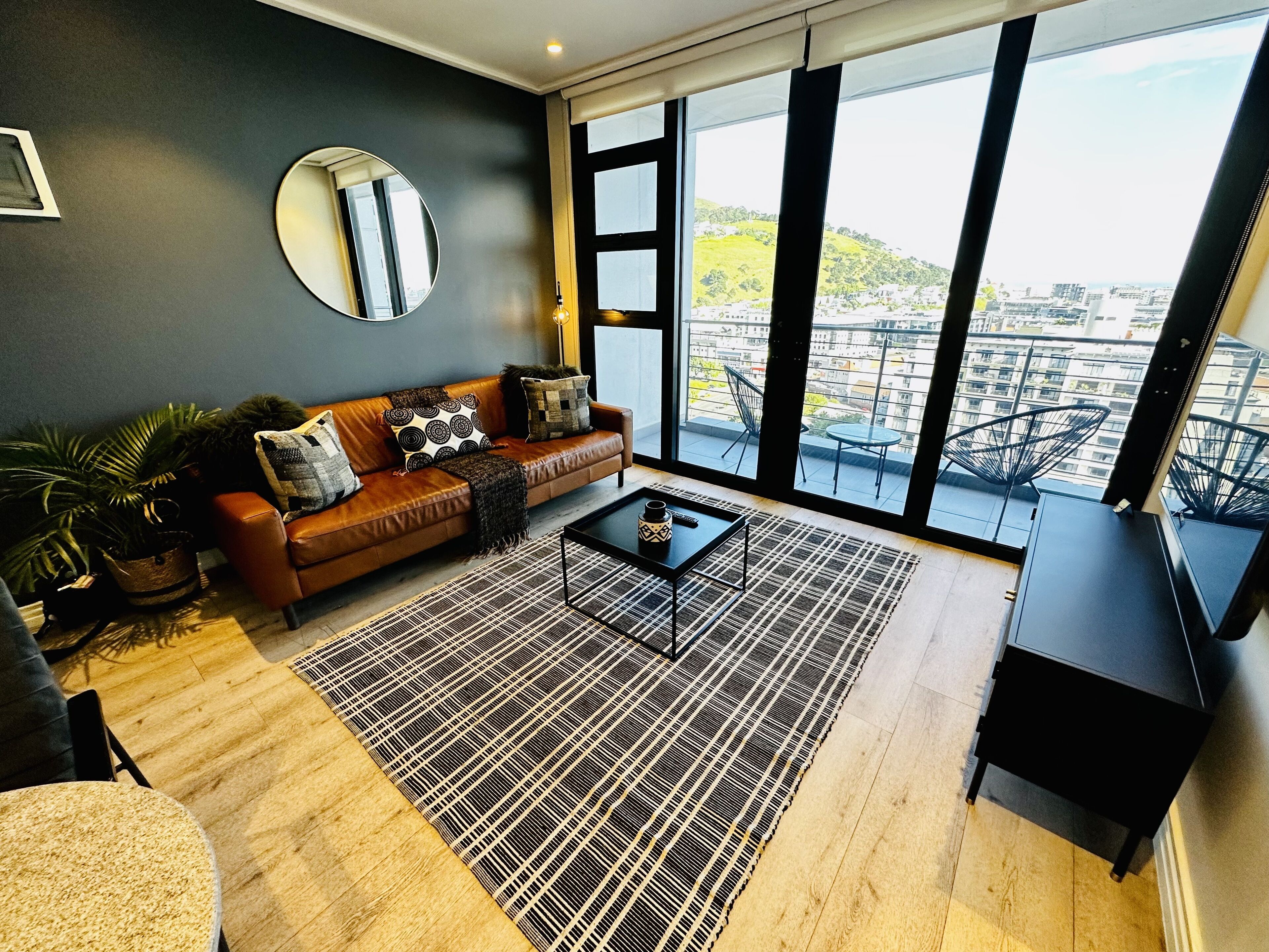 Un salón moderno con un sofá de cuero, un espejo redondo, una mesita de centro y un ventanal con vistas a la ciudad.