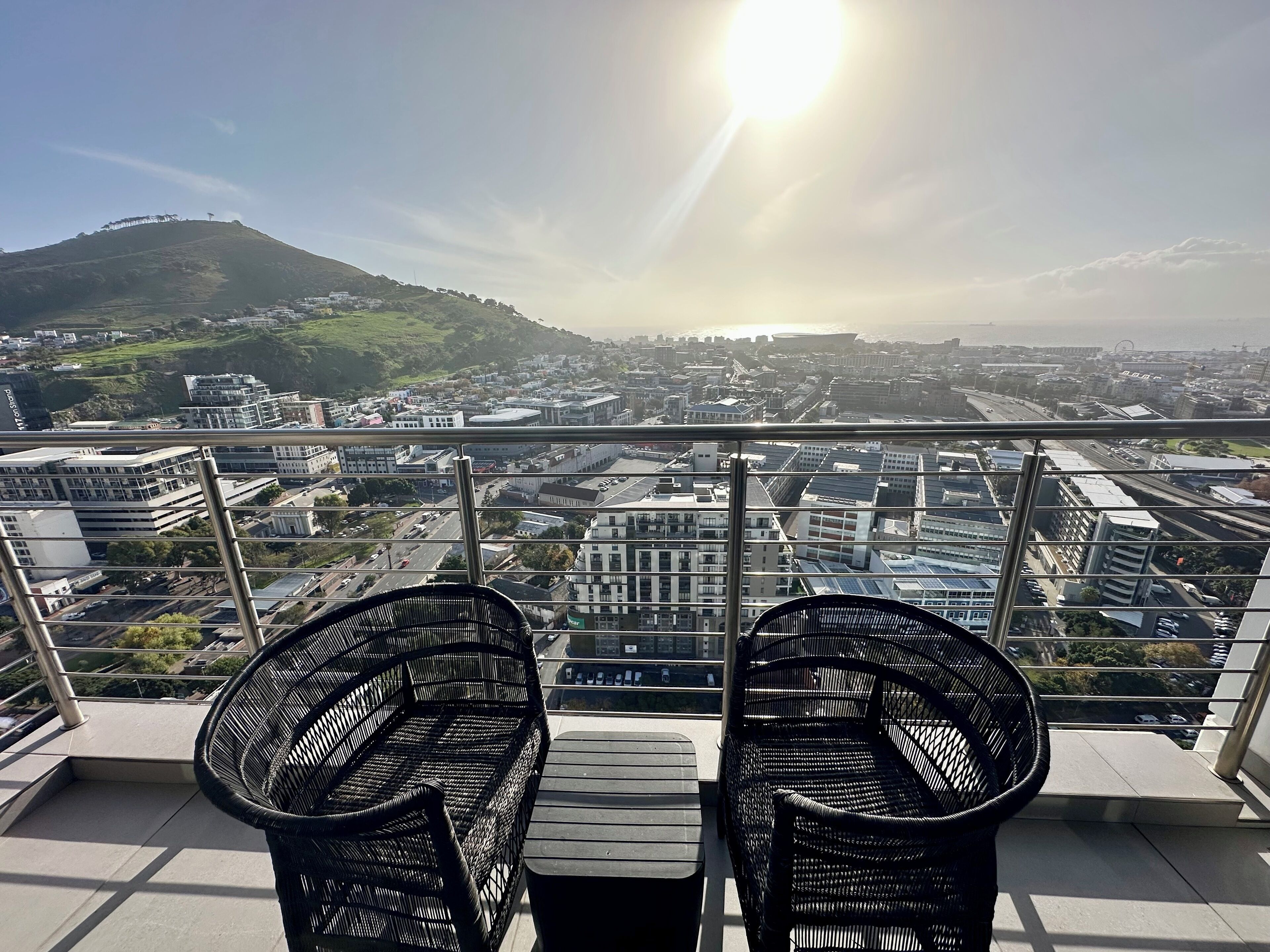 A balcony with two wicker chairs and a table, overlooking a cityscape with buildings and a hill.