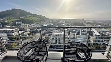 A balcony with two wicker chairs and a table, overlooking a cityscape with buildings and a hill.