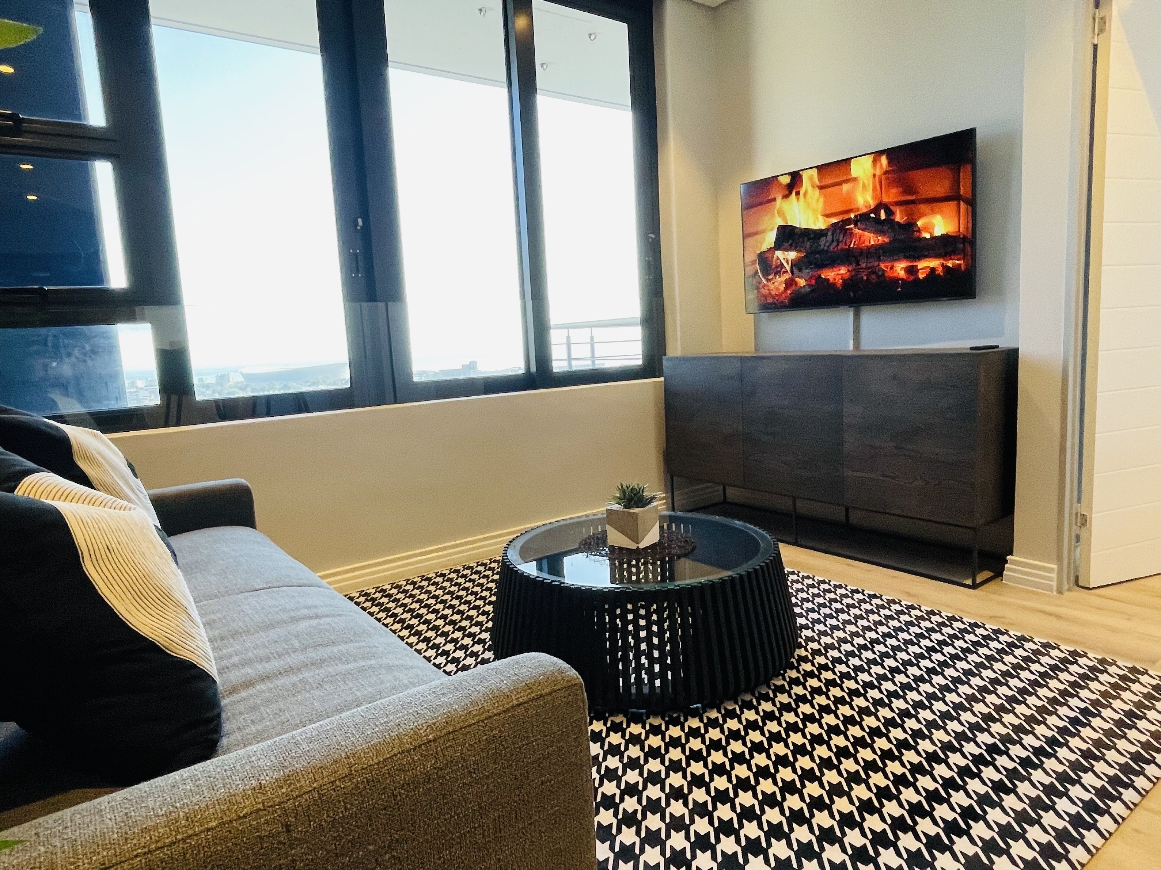A modern living room with a sofa, a coffee table with a plant, a patterned rug, a fireplace, and a large window with a city view.