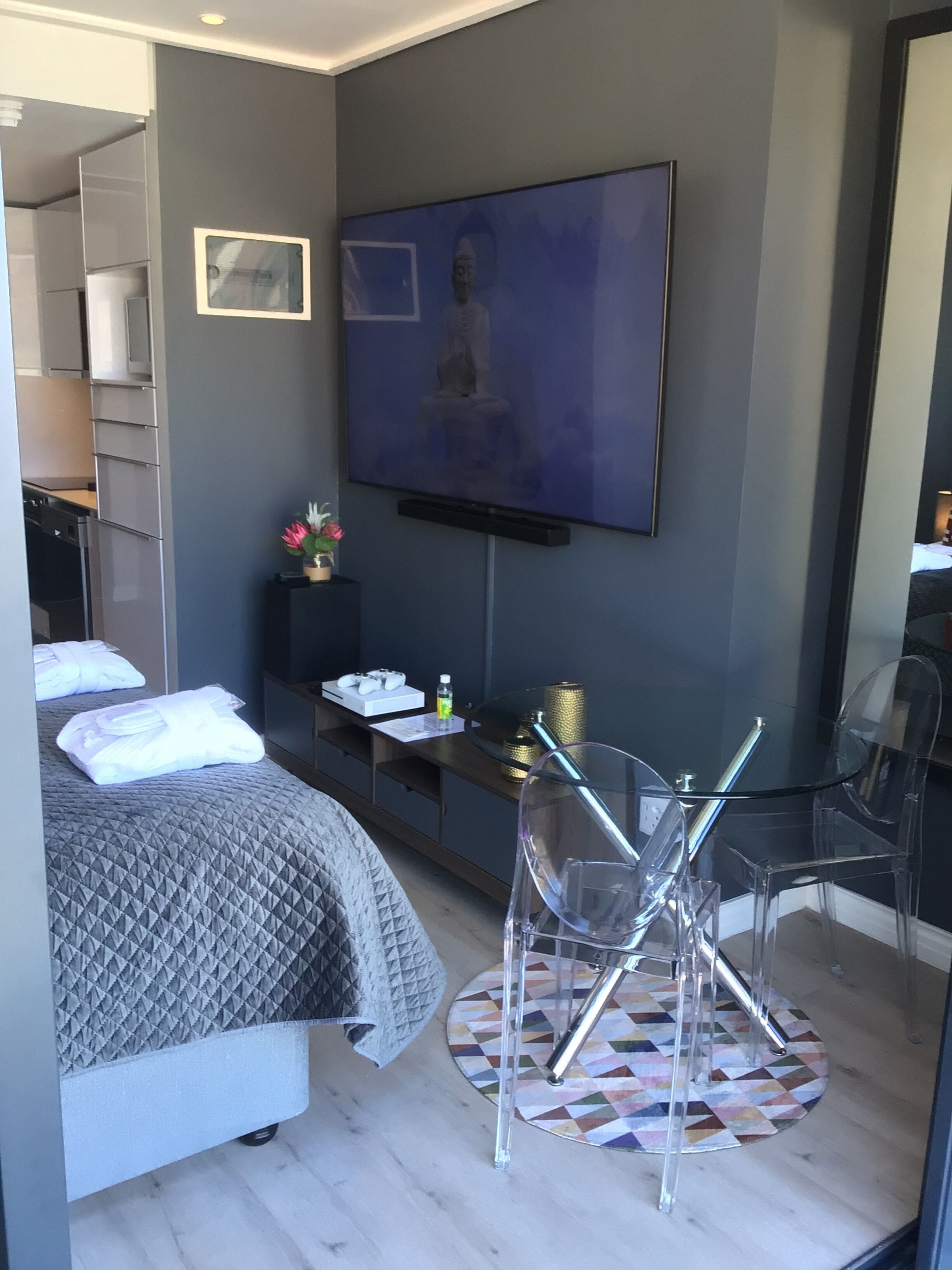 A modern hotel room with a bed, a TV mounted on the wall, a small table with a vase of flowers, and a glass chair.