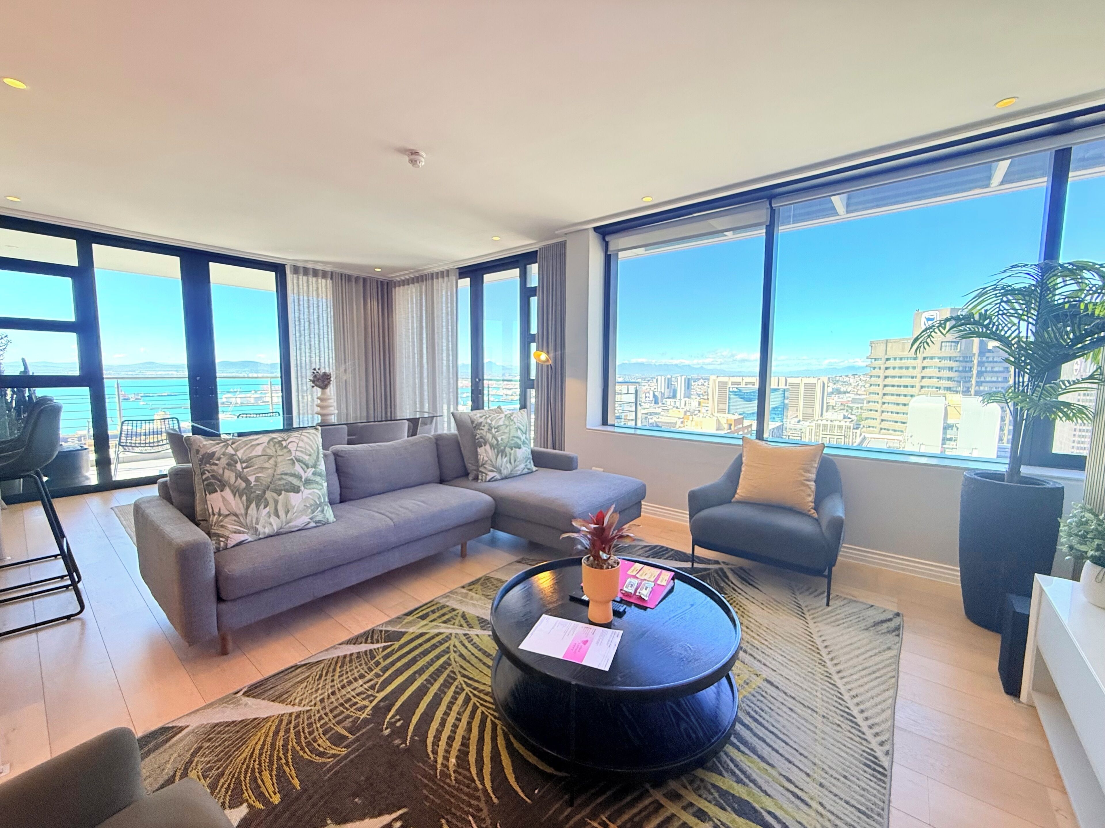 A modern living room with a sofa, armchairs, a coffee table, and large windows offering a city view.