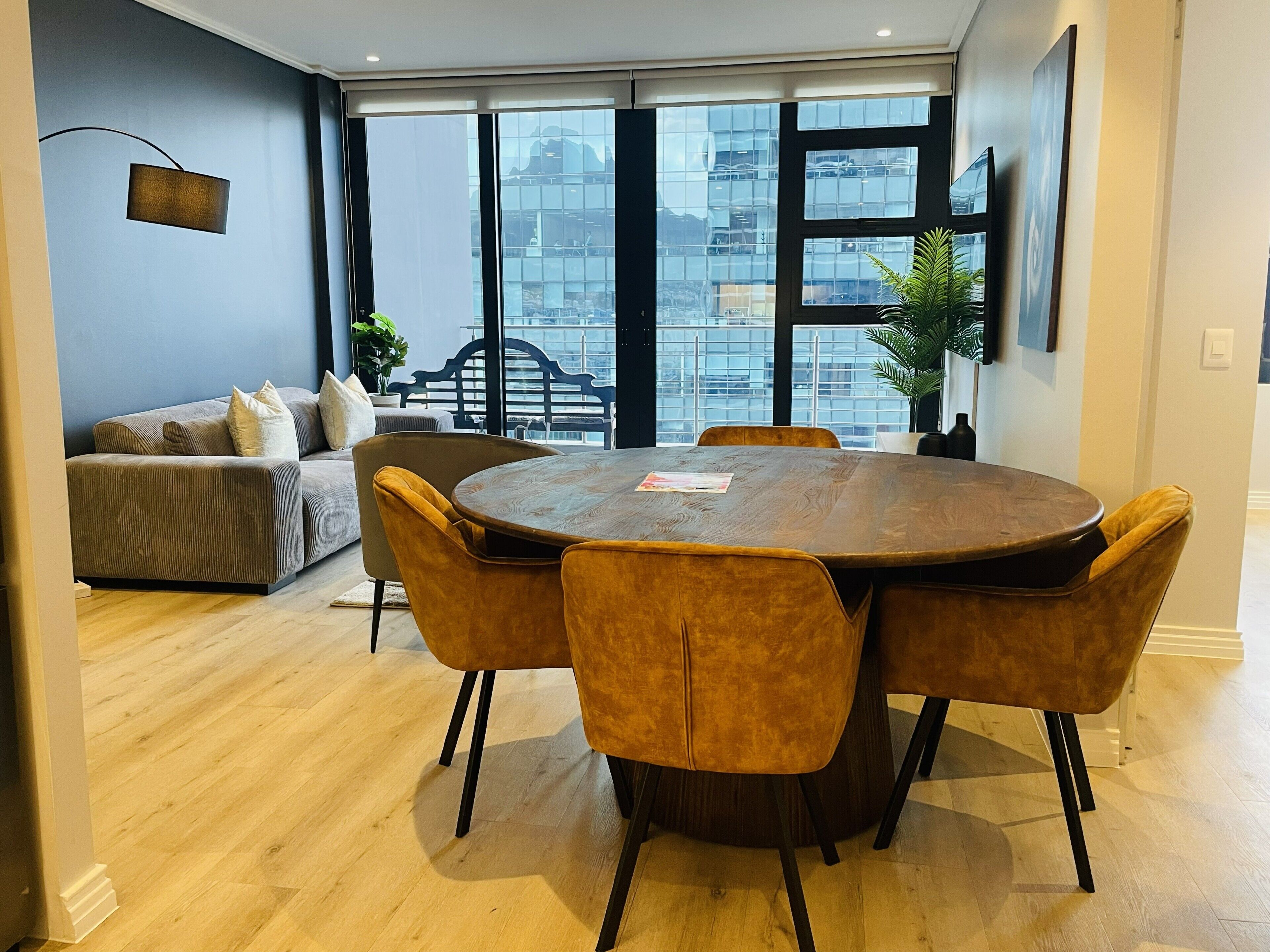 A modern living room with a round wooden table and four chairs, a grey sofa with cushions, and a large window with a view of buildings.