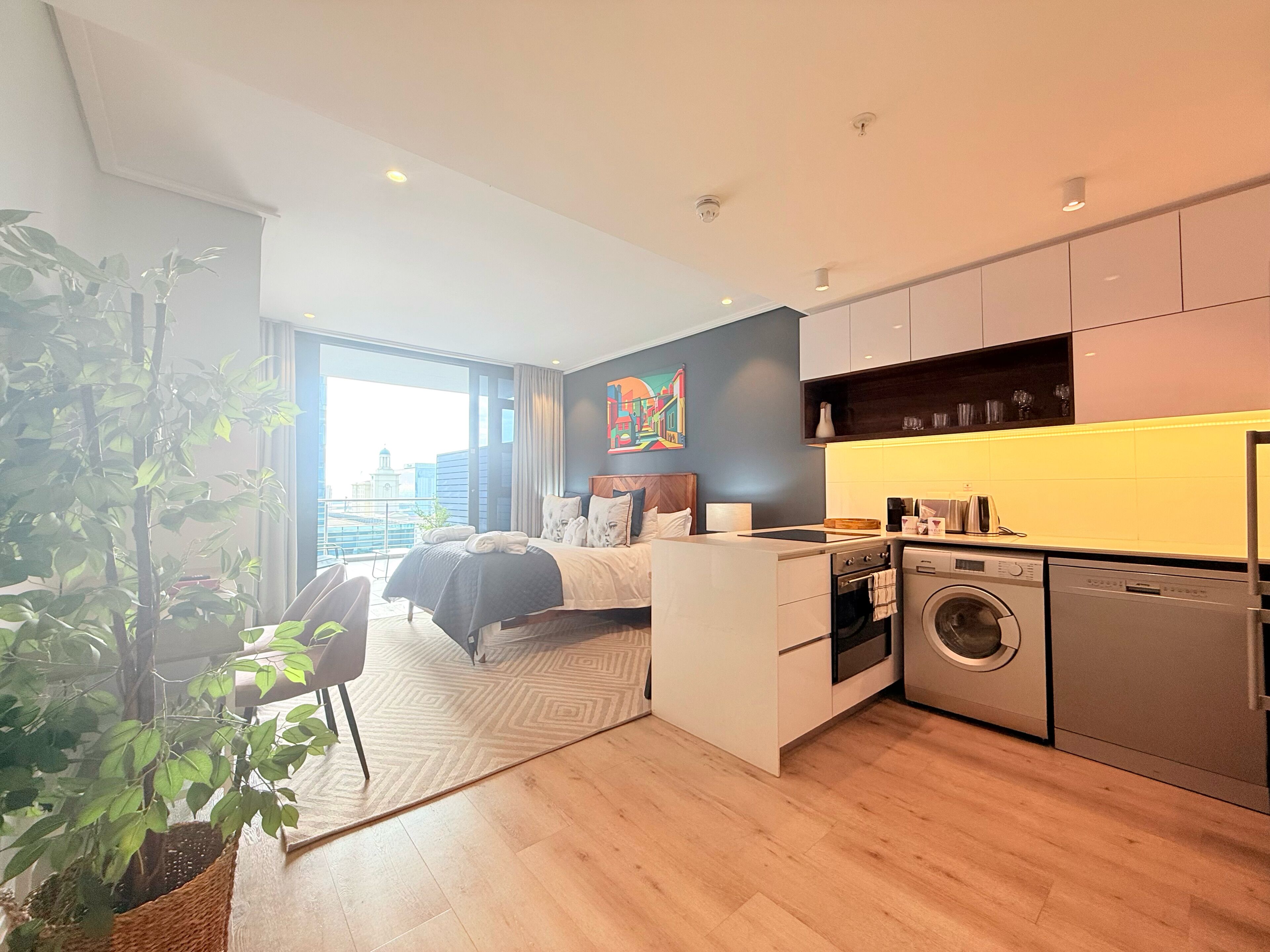 Een moderne hotelkamer met een bed, een kleine kitchenette, een wasmachine en een groot raam met uitzicht op de stad.