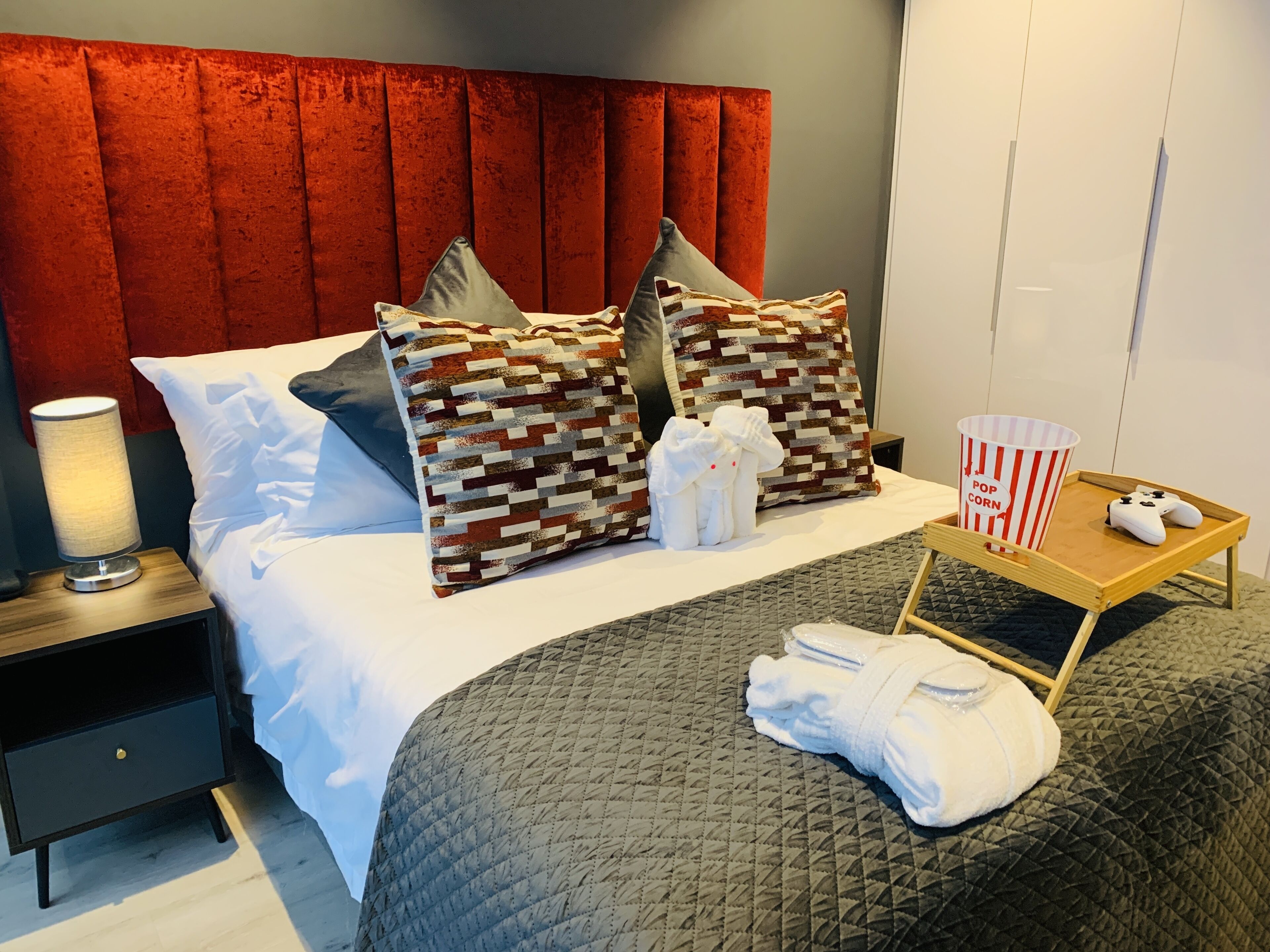 Camera d'albergo con un letto, cuscini, un comodino con una lampada, un vassoio con un contenitore di popcorn e un joystick, e un asciugamano piegato.
