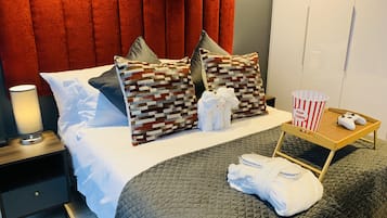Camera d'albergo con un letto, cuscini, un comodino con una lampada, un vassoio con un contenitore di popcorn e un joystick, e un asciugamano piegato.