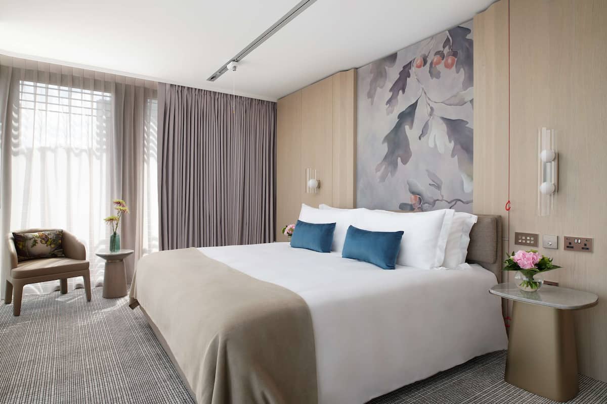 A hotel room with a large bed, a seating area with a chair, a small table with a vase, and a wall with a floral mural.