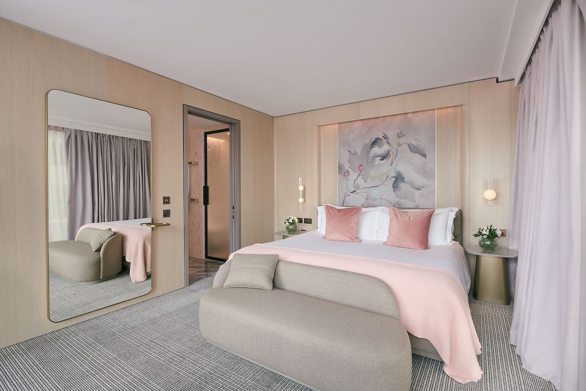 A modern hotel room with a large bed, a seating area, a mirror, and a painting on the wall.
