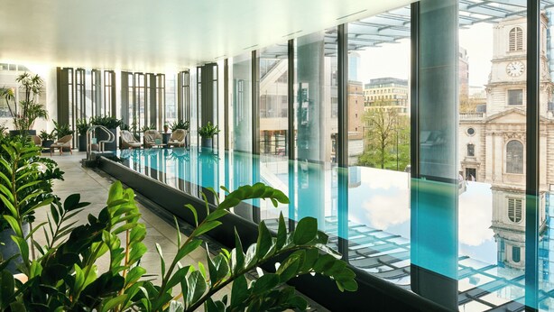A modern indoor swimming pool with large windows, potted plants, and a view of historic buildings. - Pan Pacific London (London)