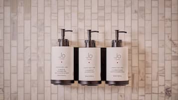 Three bottles of Jo Loves shampoo and conditioner on a tiled wall.