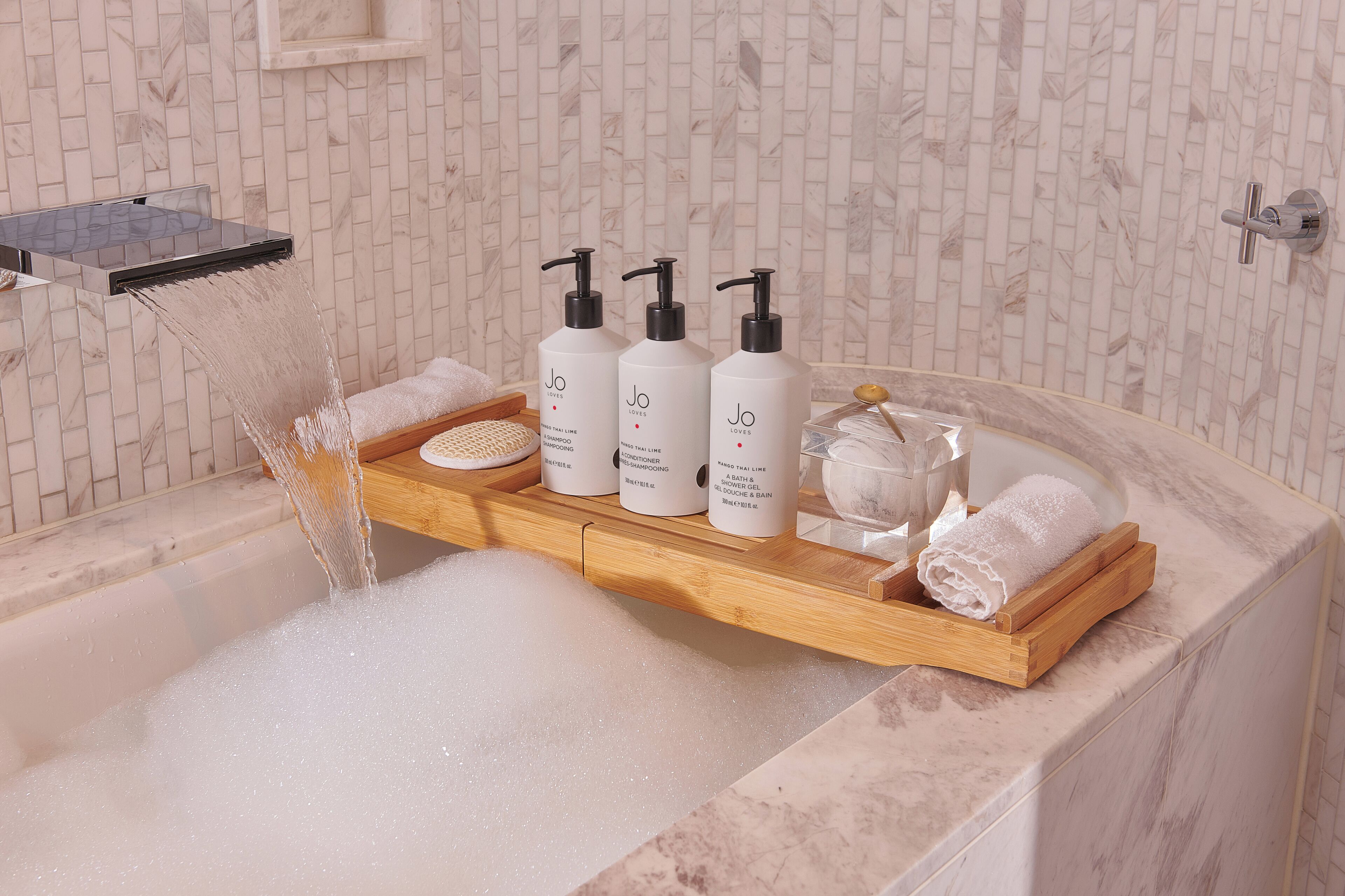 A bathtub with a wooden tray holding three bottles of shampoo and conditioner, a glass of water, and a rolled towel.