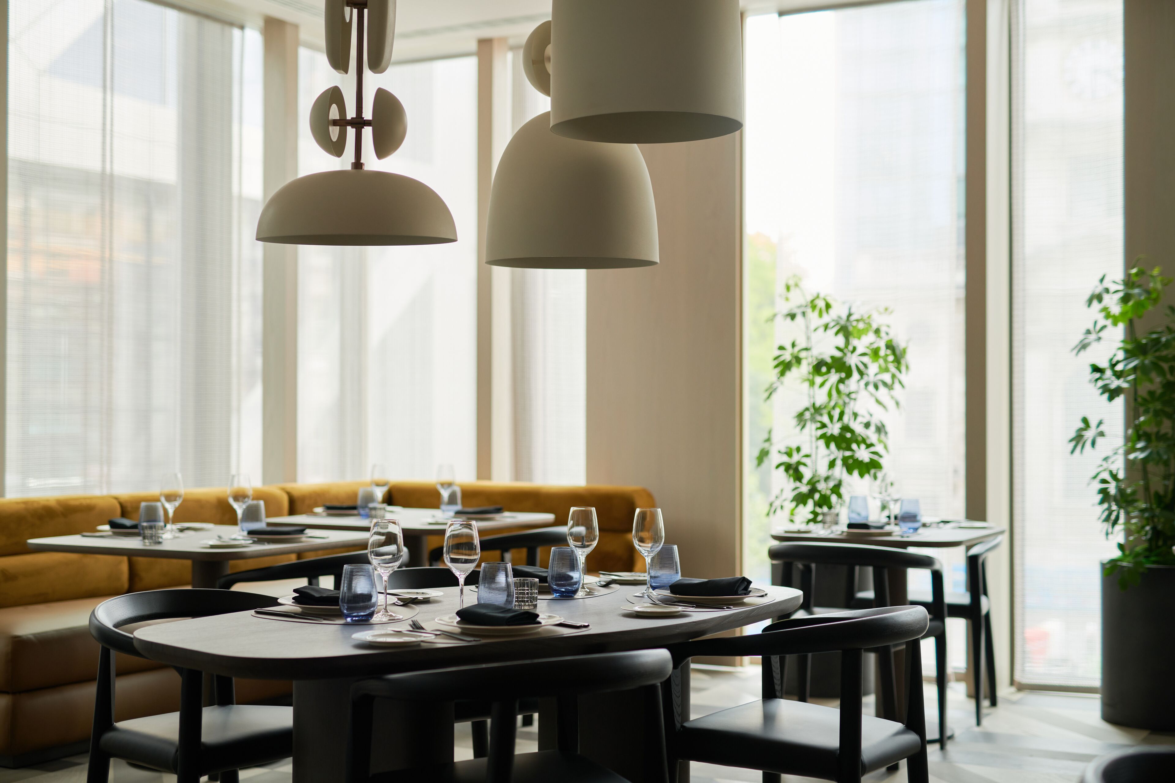 A restaurant with round tables, black chairs, and hanging pendant lights. There is a yellow sofa in the background and a potted plant on the right.