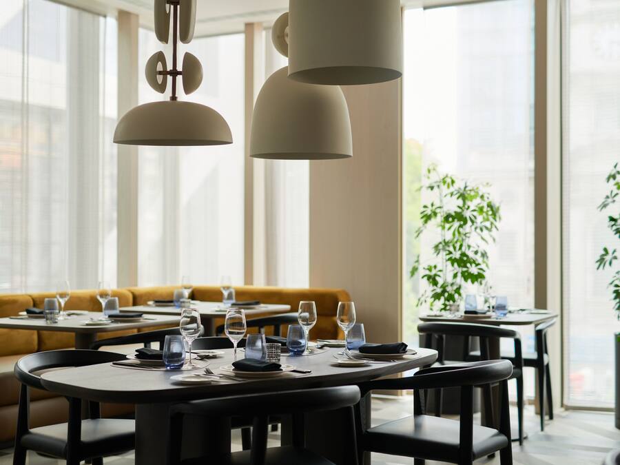 A restaurant with round tables, black chairs, and hanging pendant lights. There is a yellow sofa in the background and a potted plant on the right.
