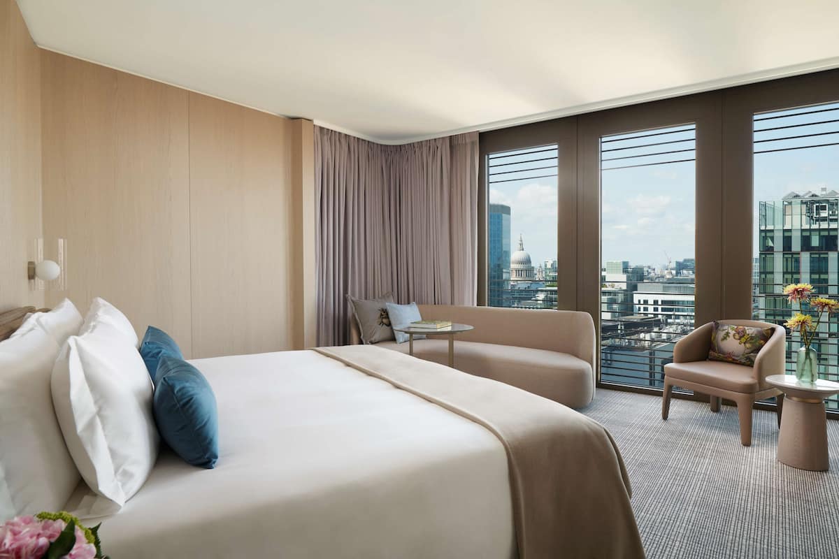 A hotel room with a large bed, a sofa, a small table, and a view of the cityscape through large windows.