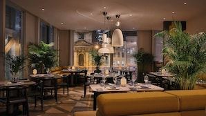 A restaurant with multiple dining tables and chairs, large windows, and potted plants. - Pan Pacific London (London)