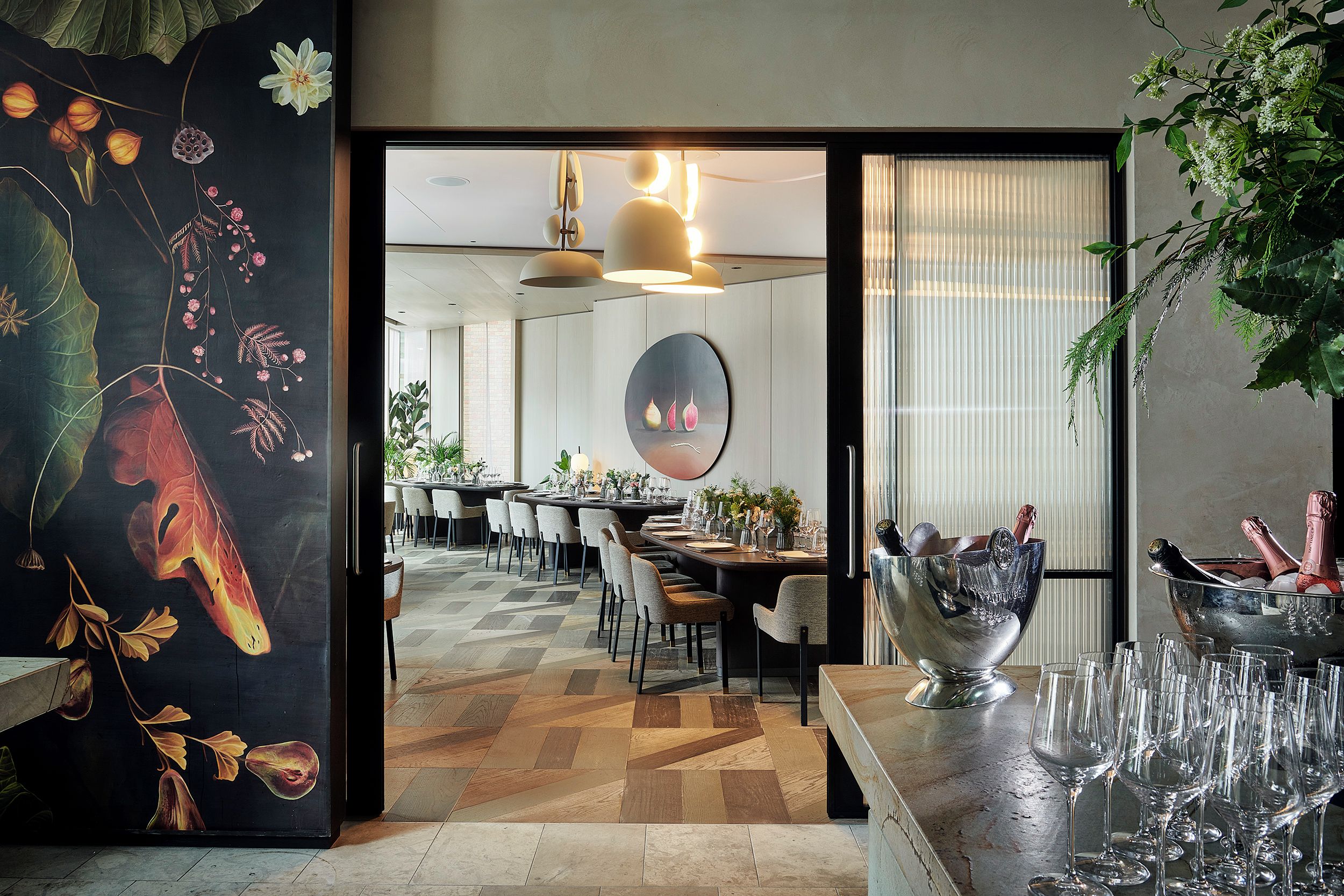 A modern dining area with a large table set for a meal, a decorative wall with a fruit and flower painting, and a large window with a view.