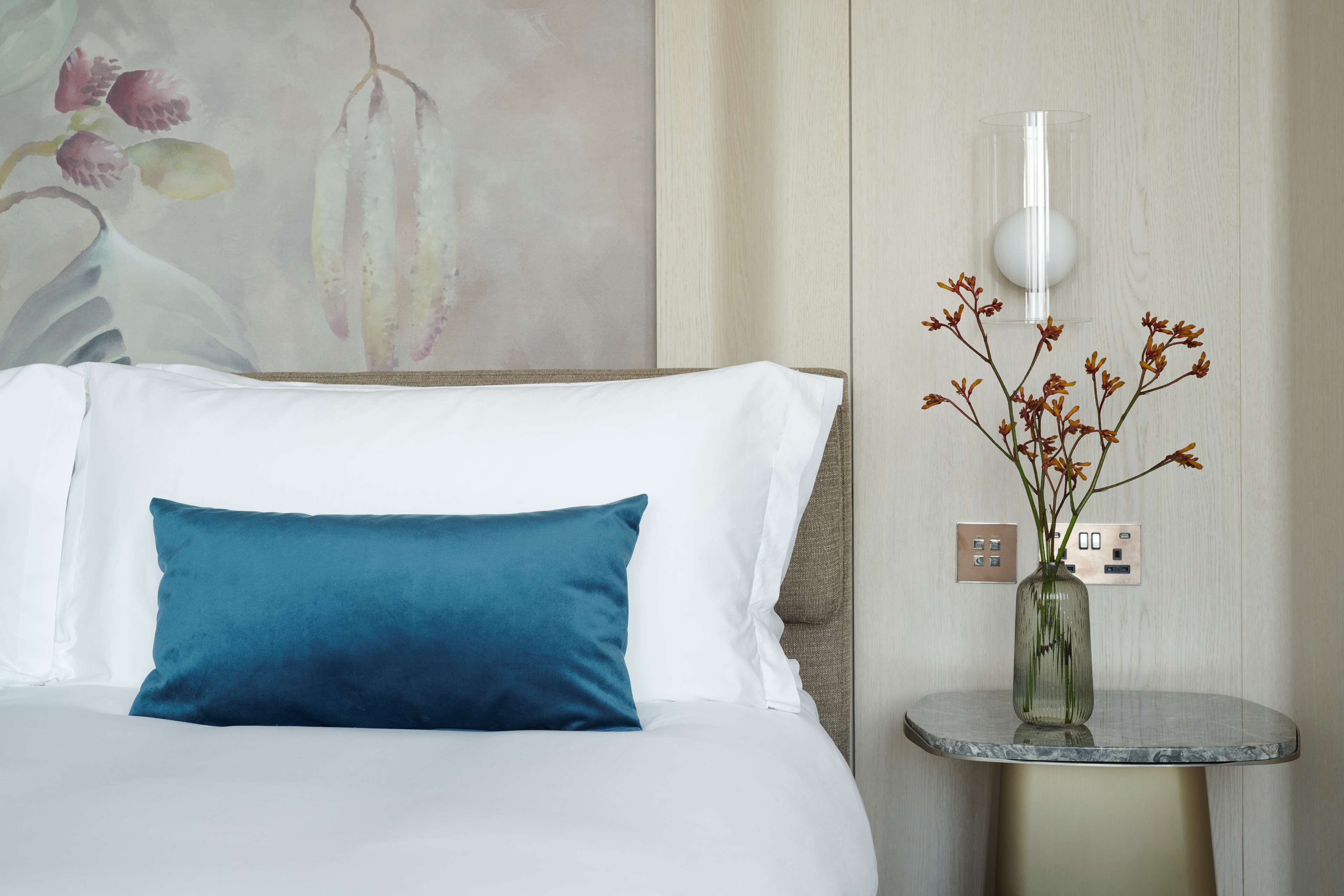 A neatly made bed with white and blue pillows, a vase with flowers on a small table beside it, and a wall-mounted light fixture.