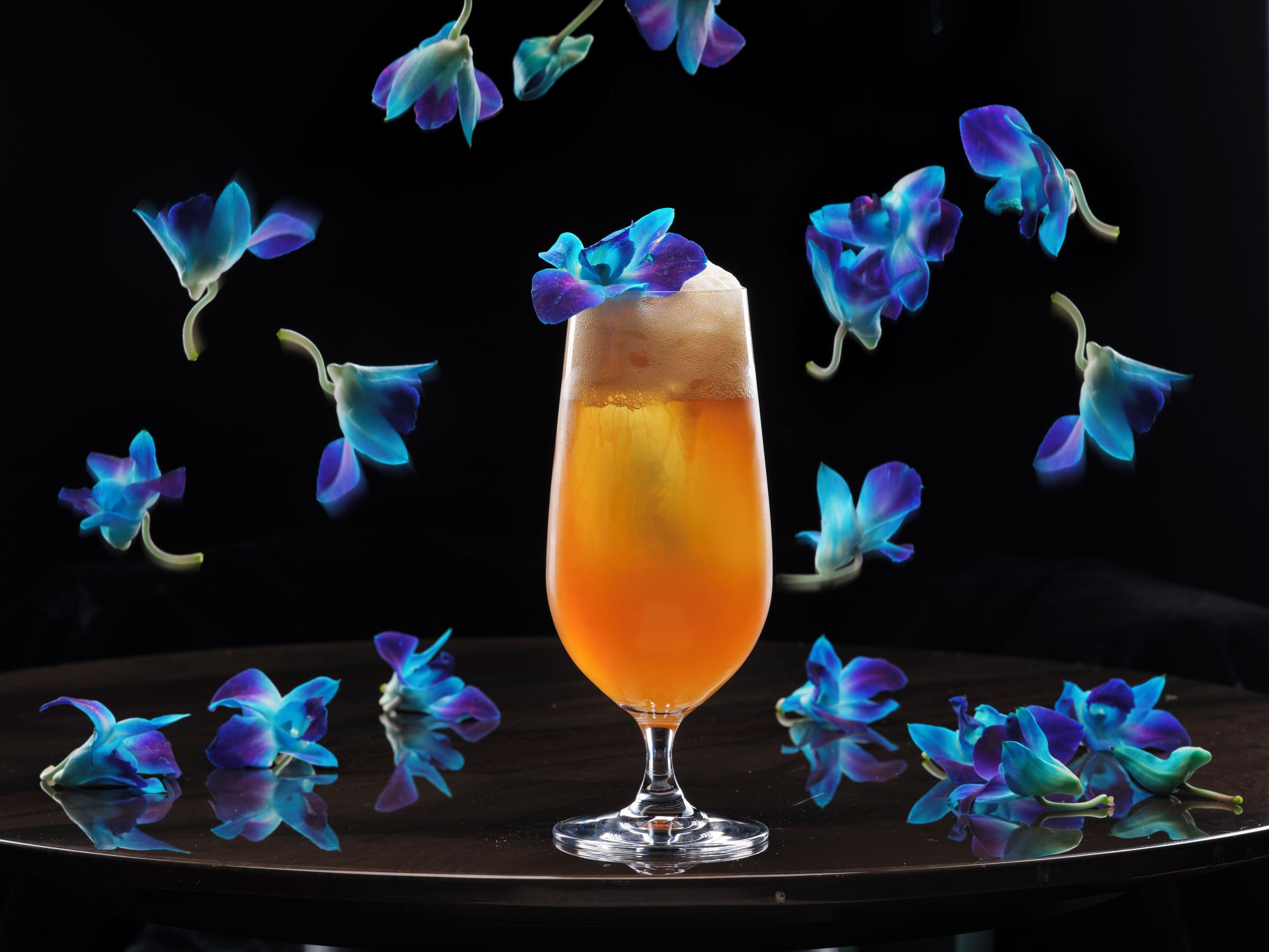 A glass of orange liquid on a reflective surface with blue flowers floating around.
