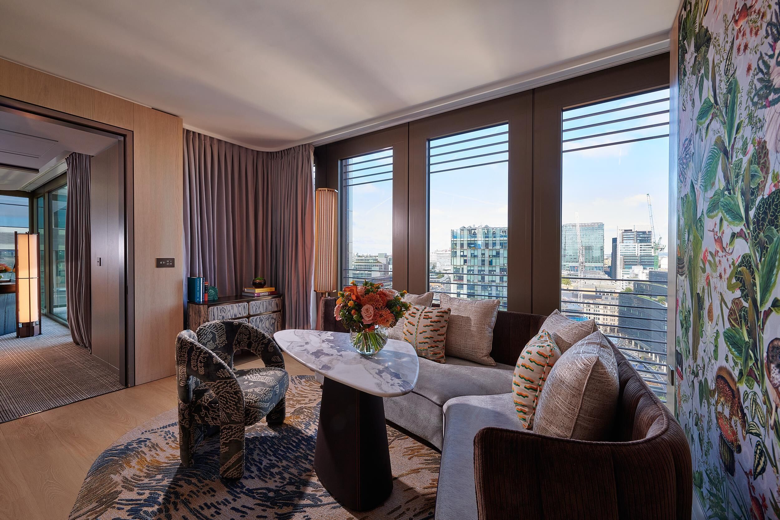 A modern hotel room with a large window, a sofa, a patterned armchair, and a coffee table with a vase of flowers.