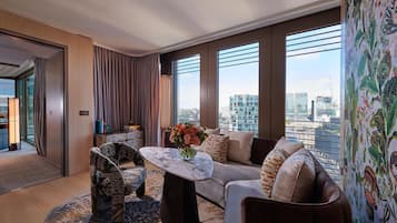 A modern hotel room with a large window, a sofa, a patterned armchair, and a coffee table with a vase of flowers.