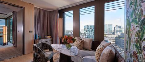 Una habitación de hotel moderna con un ventanal grande, un sofá, un sillón estampado y una mesa de centro con un florero.