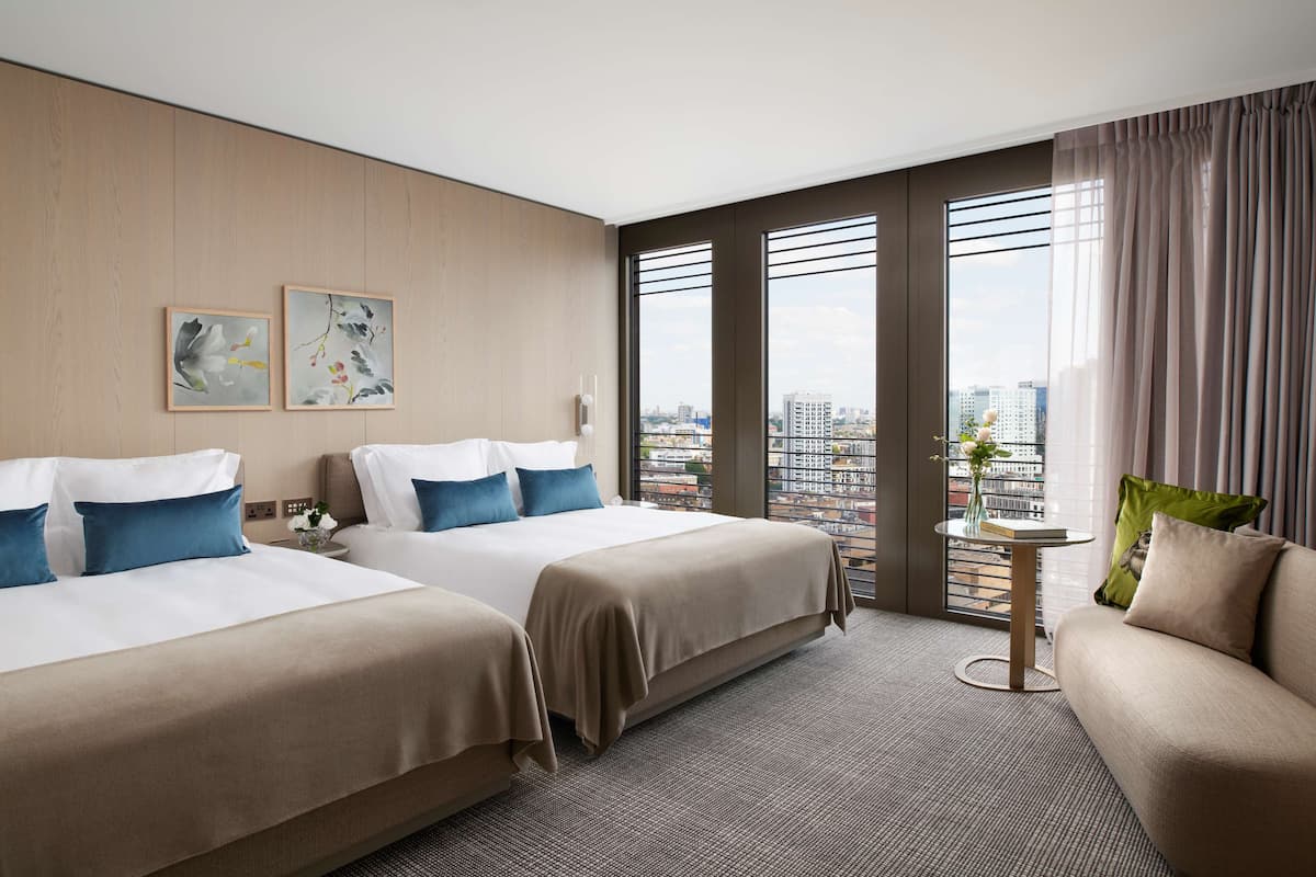 A hotel room with two beds, a sofa, a small table with a vase, and large windows with a city view.