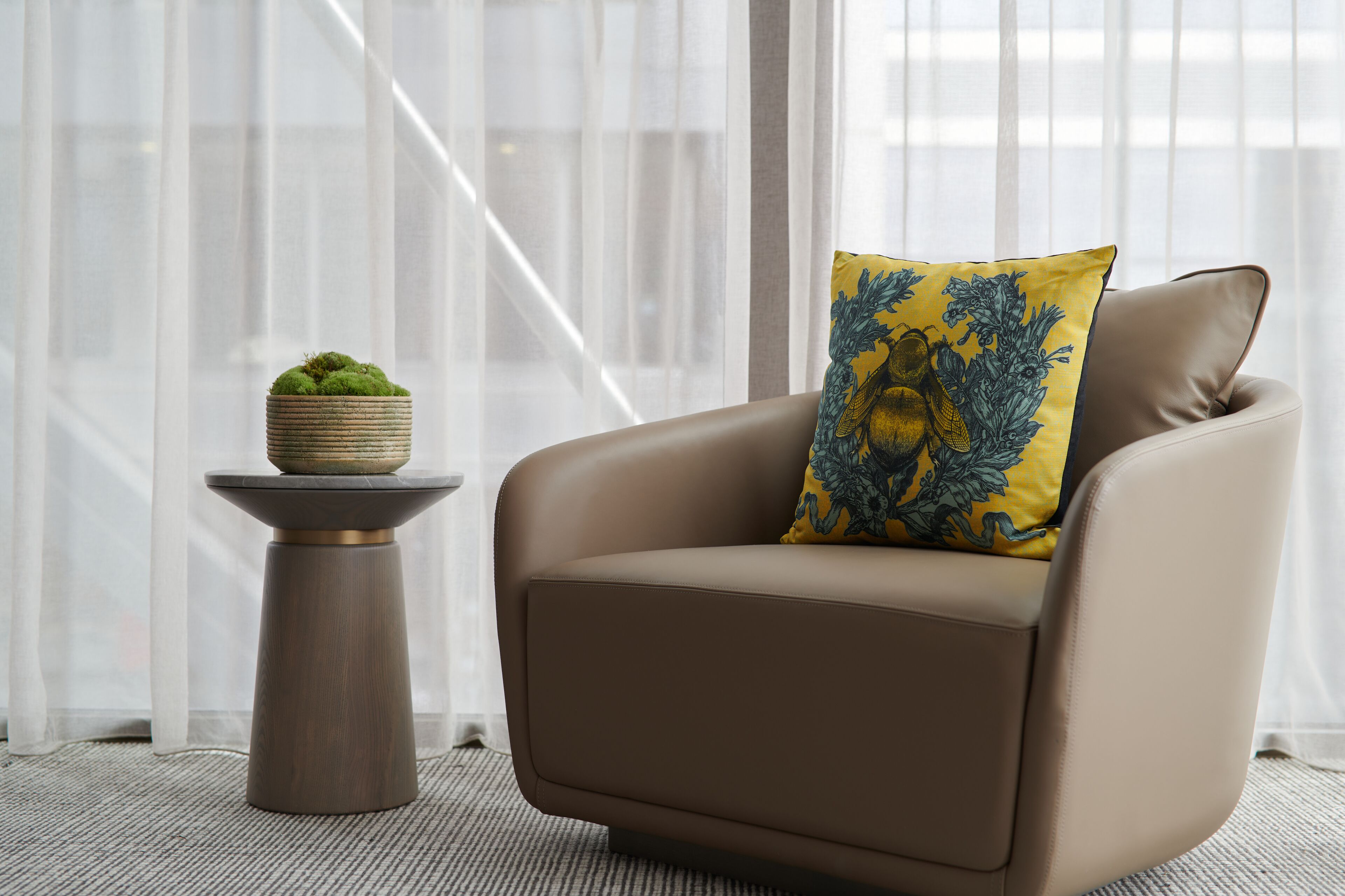 A modern armchair with a decorative pillow, a small side table with a plant, and a view of a staircase through sheer curtains.