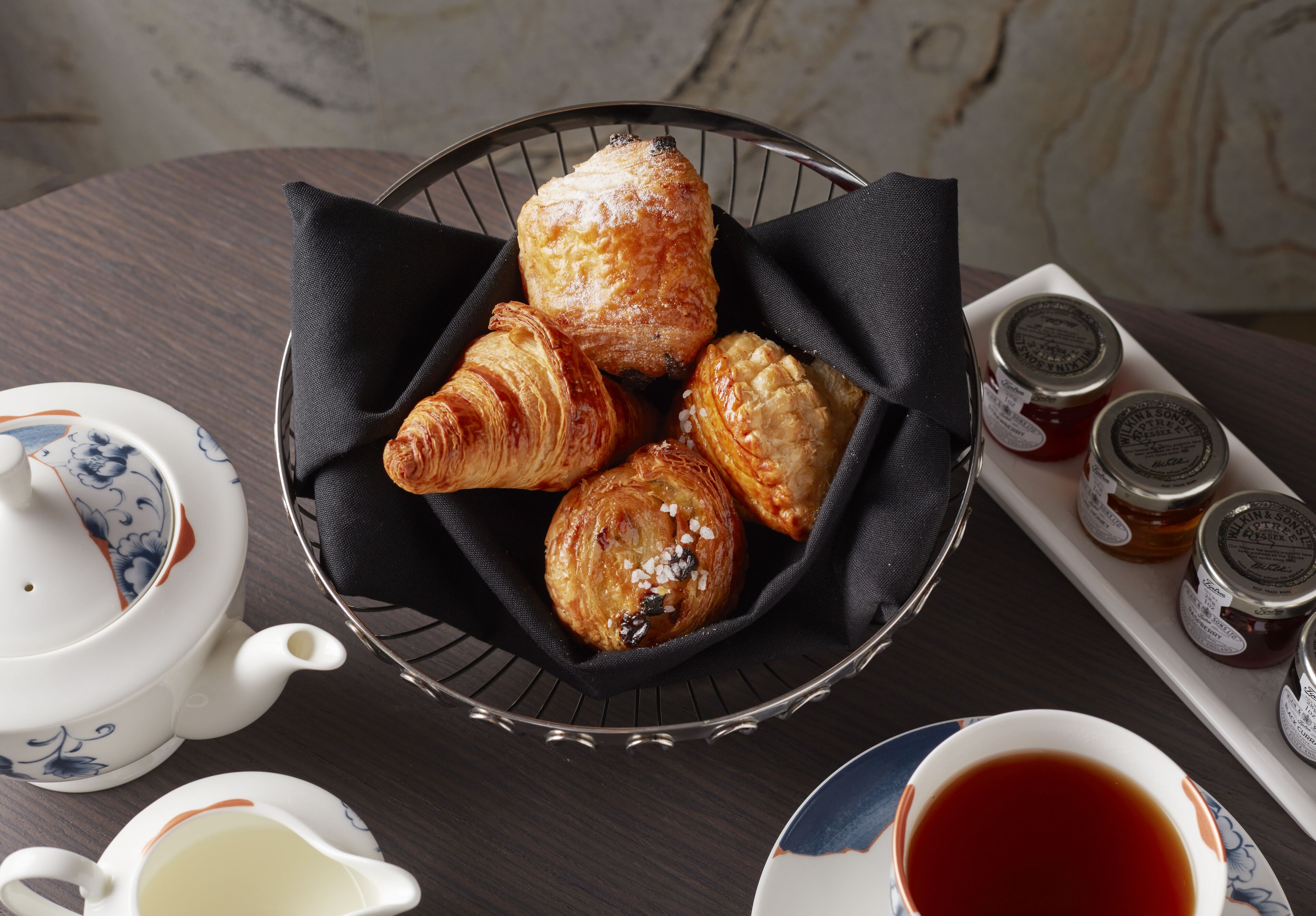 A table with a basket of pastries, a teapot, a cup of tea, and jars of jam.