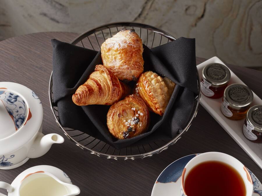 A table with a basket of pastries, a teapot, a cup of tea, and jars of jam.