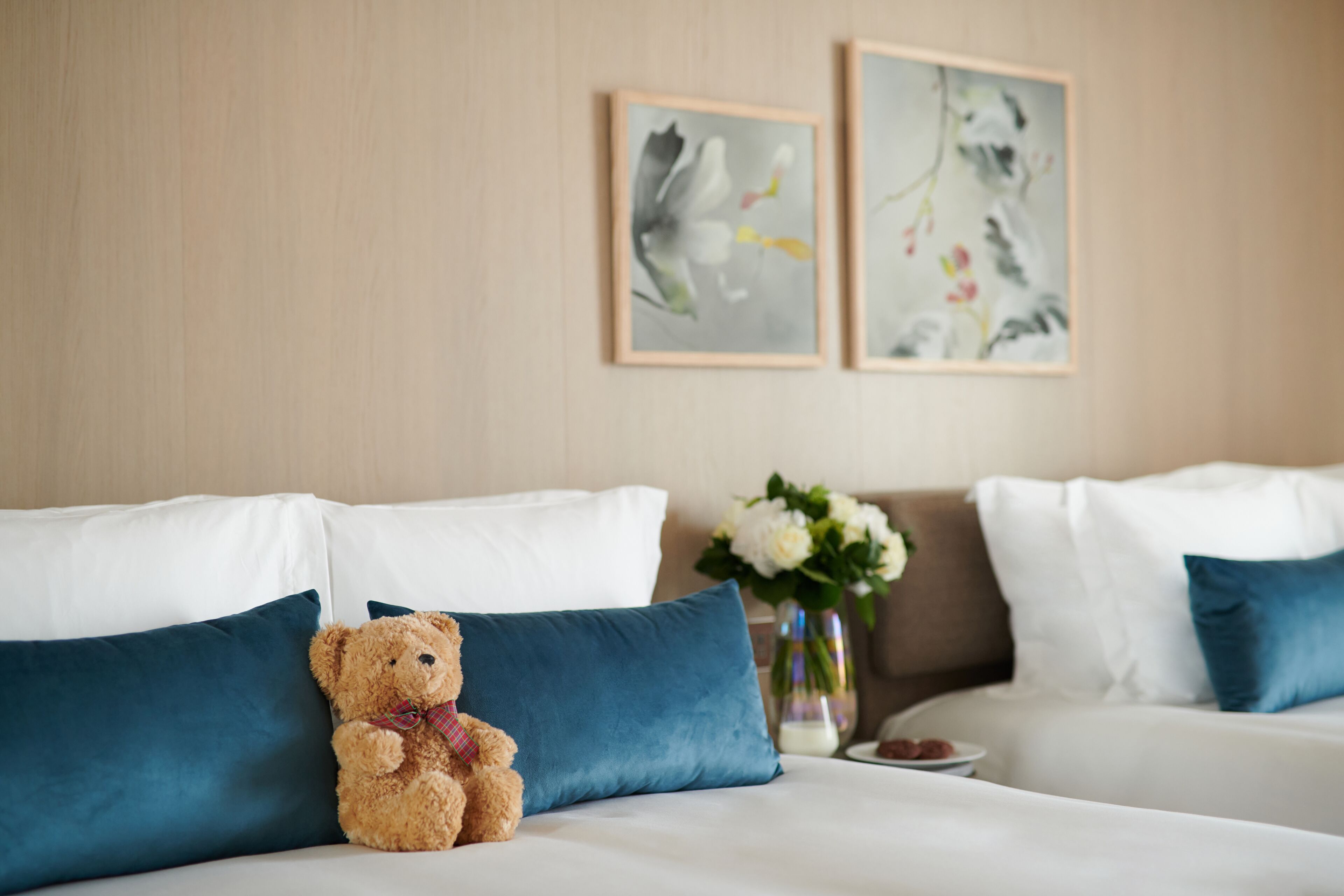 Habitación de hotel con dos camas, cada una con ropa de cama blanca y almohadas azules. Un oso de peluche está sobre la cama de la izquierda. En la pared detrás de las camas hay dos cuadros con motivos florales. Un jarrón con flores se encuentra en la parte derecha de la habitación.