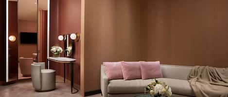 A modern hotel room with a sofa, a round table with a vase of flowers, a small round table with a book, a mirror, and a television.