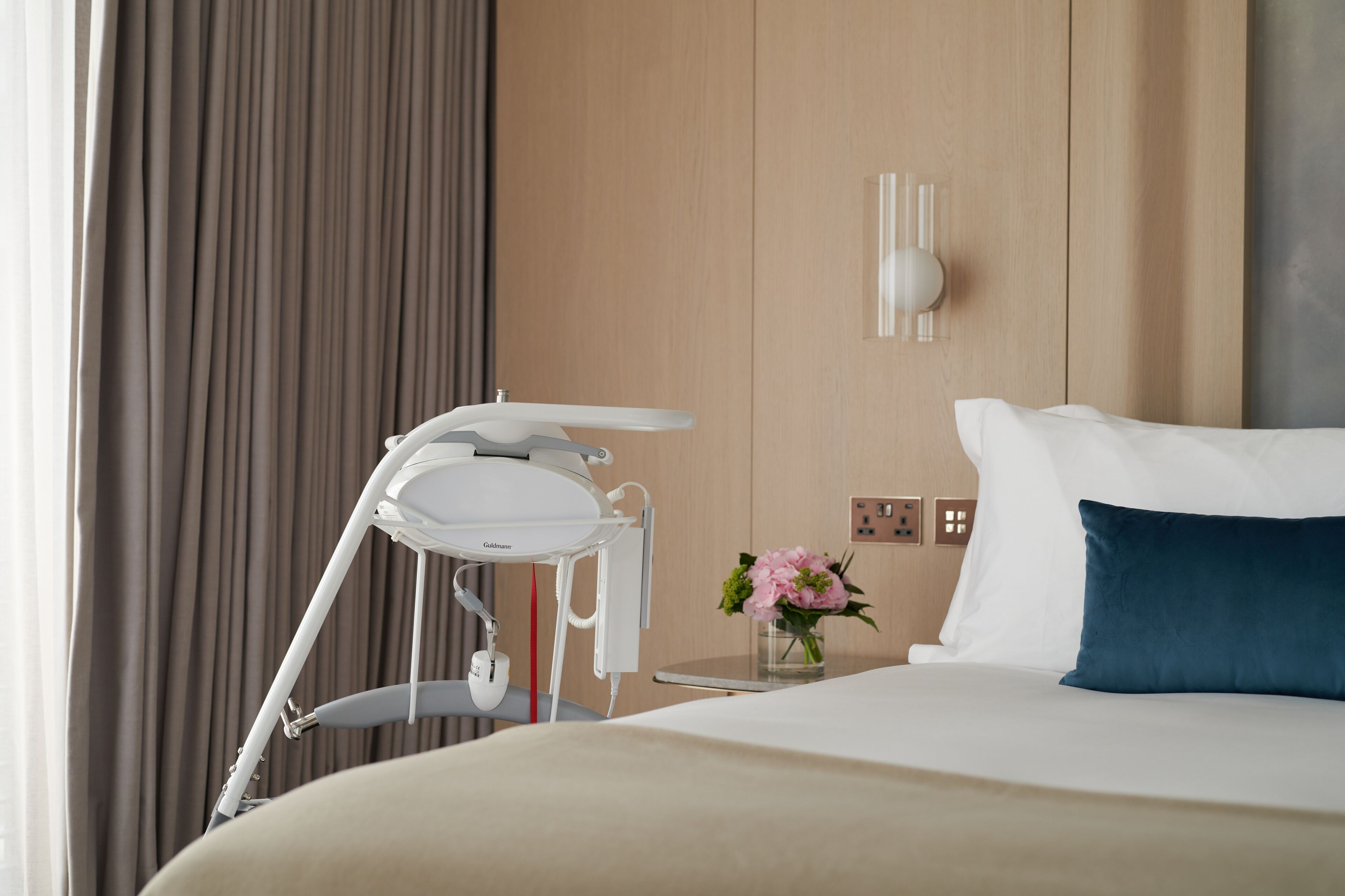 A hotel room with a bed, a bedside table with a vase of flowers, a white bedside lamp, and a white chair.