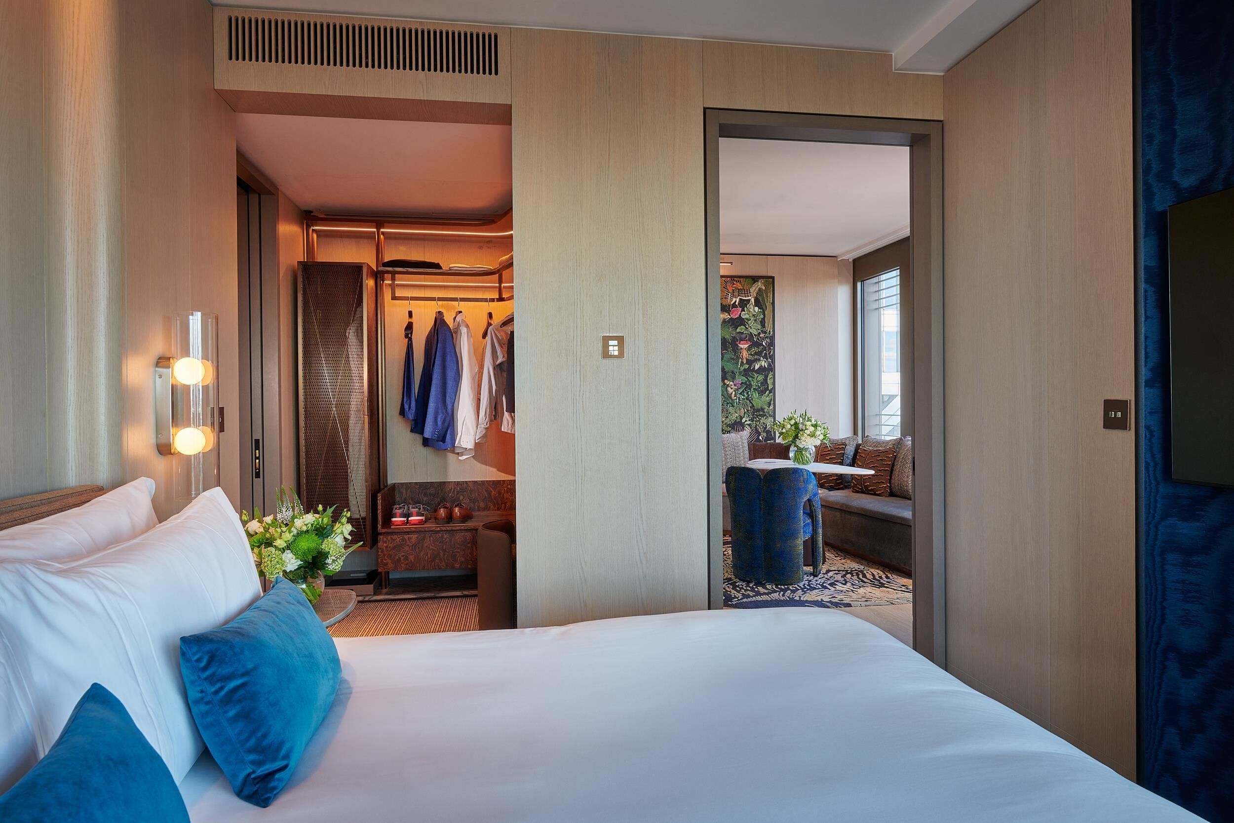 A hotel room with a large bed, a sitting area with a sofa and a small table, a bathroom with a shower and a mirror, and a view of the outside through a large window.