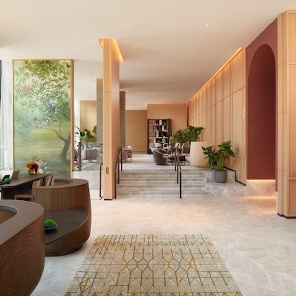 A modern hotel lobby with a curved reception desk, a staircase leading to the upper floor, and a large painting on the wall.