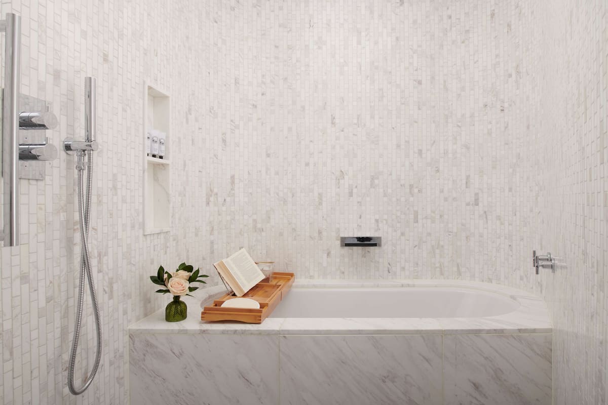 A modern bathroom with a white tiled wall, a bathtub, a showerhead, a small shelf with toiletries, and a wooden tray with a book and a vase of flowers.
