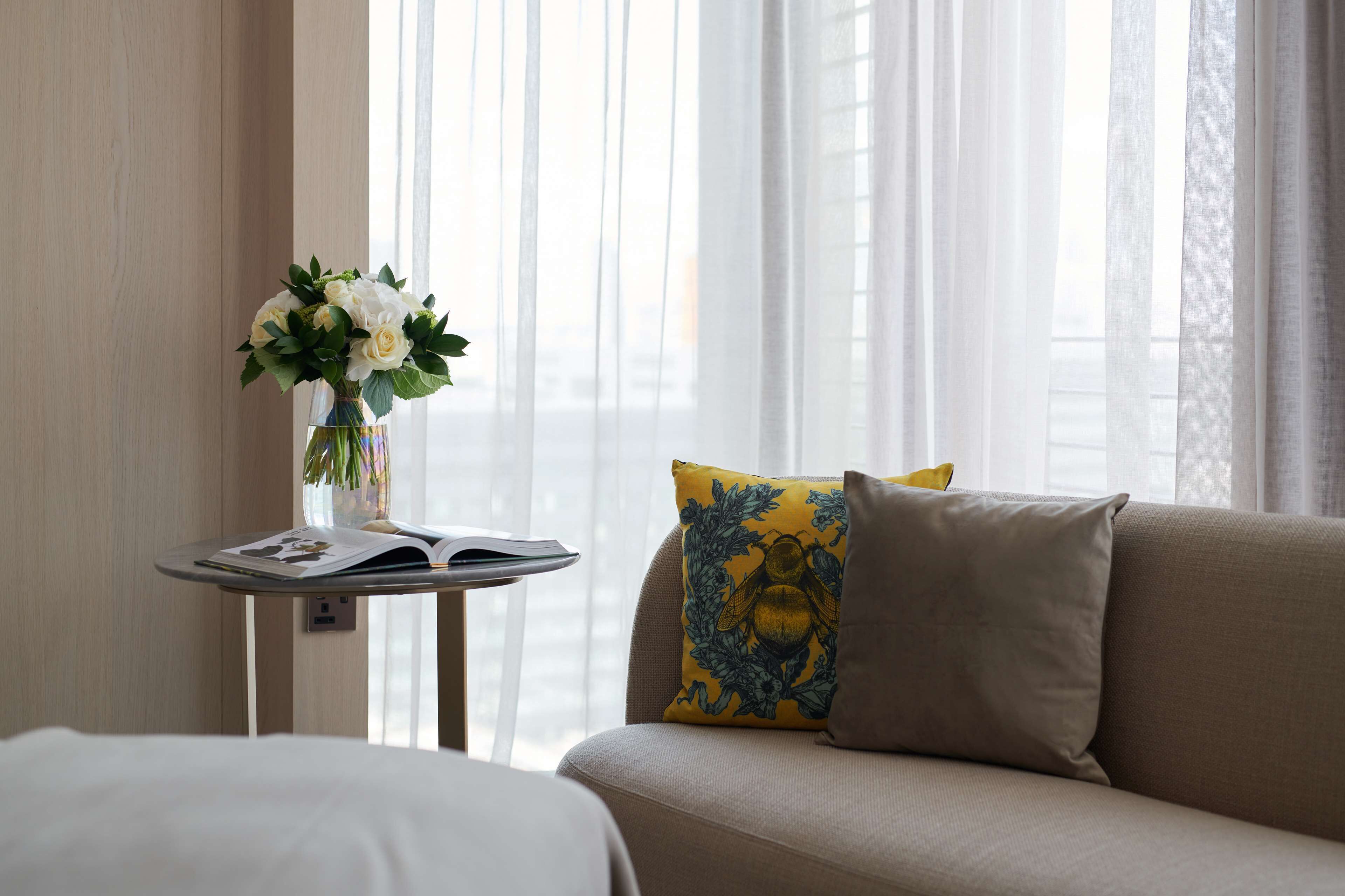 A hotel room with a sofa, a small table with a vase of flowers, and a window with sheer curtains.