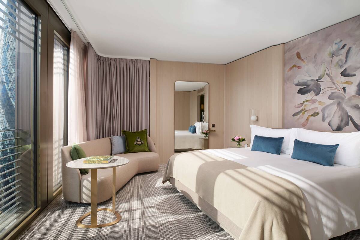 A modern hotel room with a large bed, a curved sofa, a small round table, and a large window with blinds.