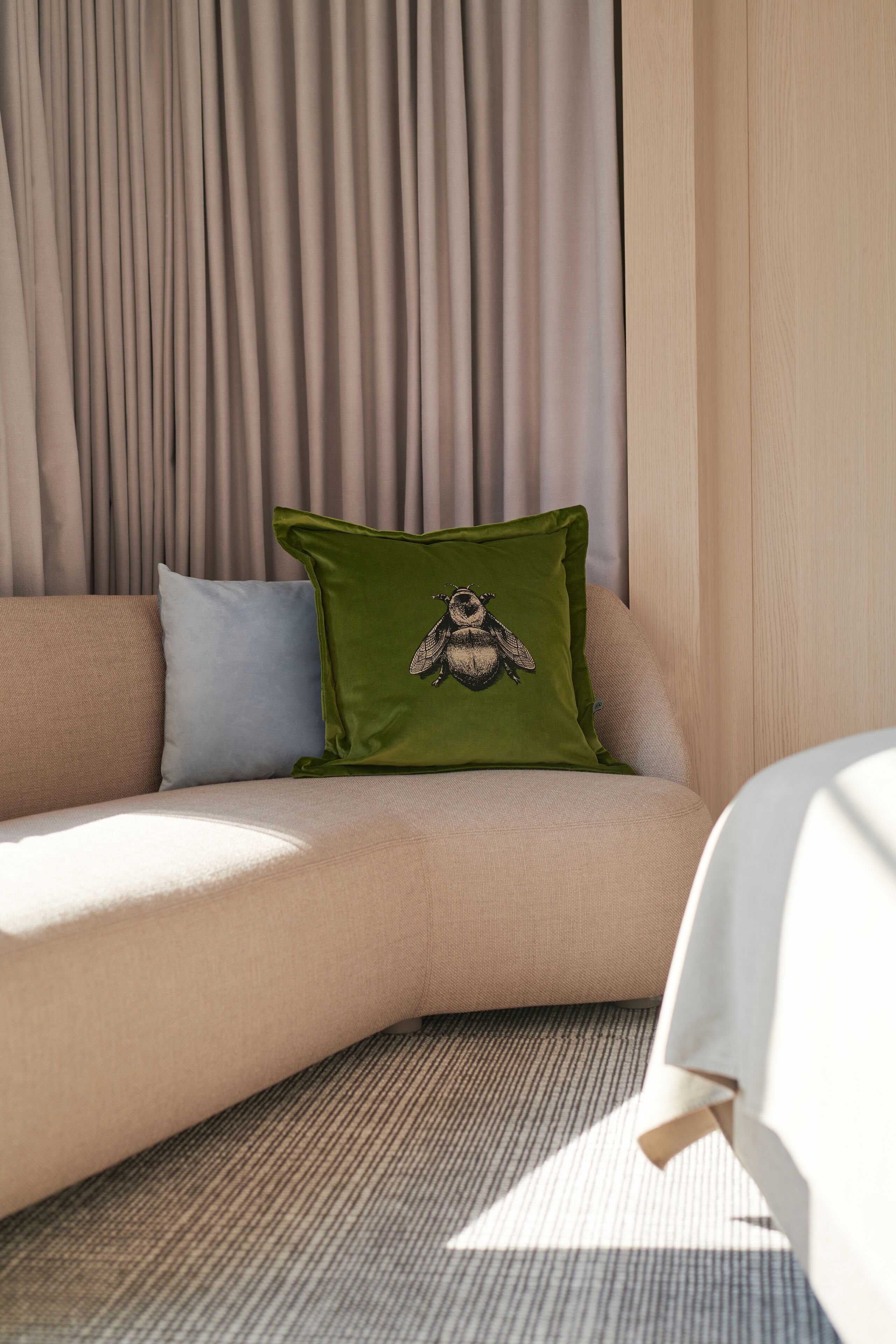 A corner of a room with a beige sofa, a green pillow with a fly design, and a light-colored wall.
