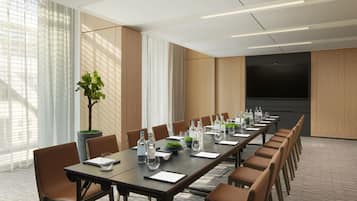 Un salón de conferencias con una larga mesa, sillas, una pantalla grande y una planta en maceta.