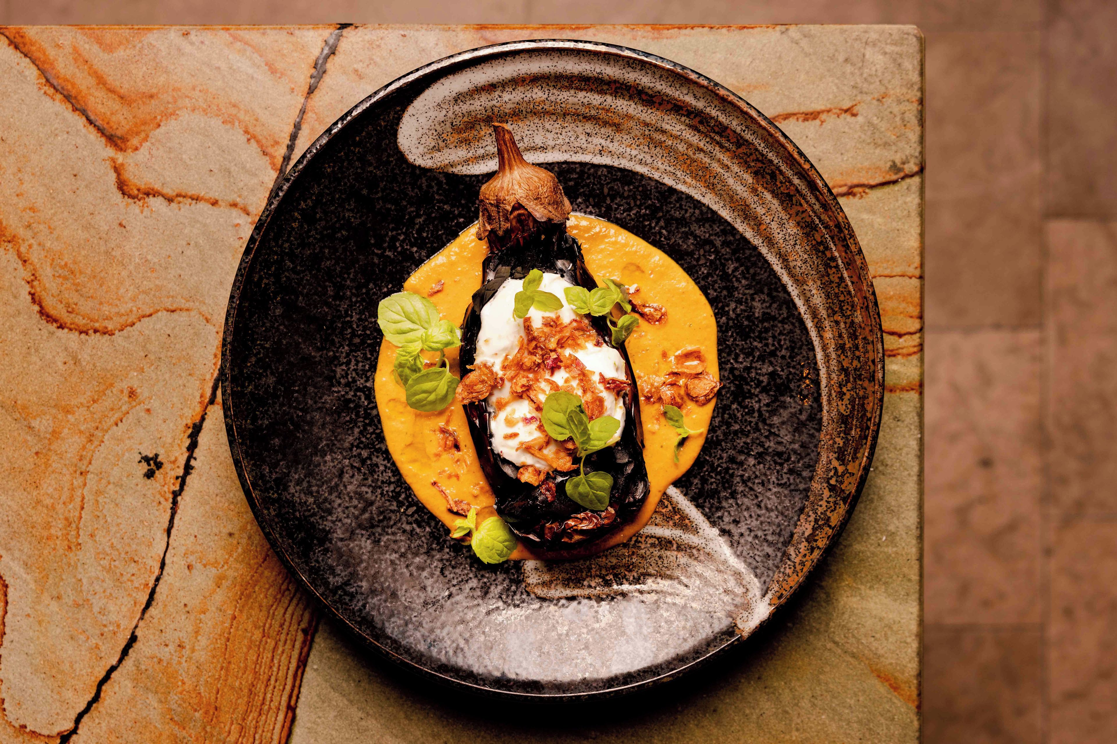 A dish with a roasted eggplant, a dollop of cream, and garnishes on a ceramic plate.