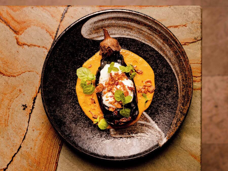 A dish with a roasted eggplant, a dollop of cream, and garnishes on a ceramic plate.